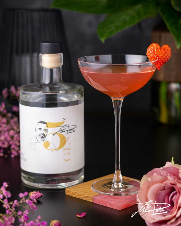Das Foto zeigt ein Cocktailglas mit Gin Cocktail Cosmo Valentine auf Holz-Harz-Untersetzer, eine Rose, Luis Dias Gin N° 5 Flasche, herzförmige Erdbeere, schwarzer Tisch, dunkler Hintergrund zum Muttertag