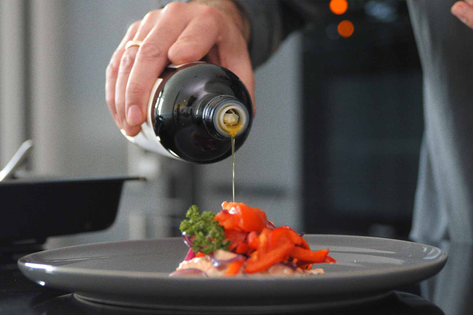 Das Foto zeigt Luis Dias' Hand, die eine Flasche Olivenöl Extra Vergine waagerecht hält und das kaltgepresste Olivenöl über ein appetitlich angerichtetes Gericht auf einem Teller gießt, um Geschmack und Qualität zu verfeinern.