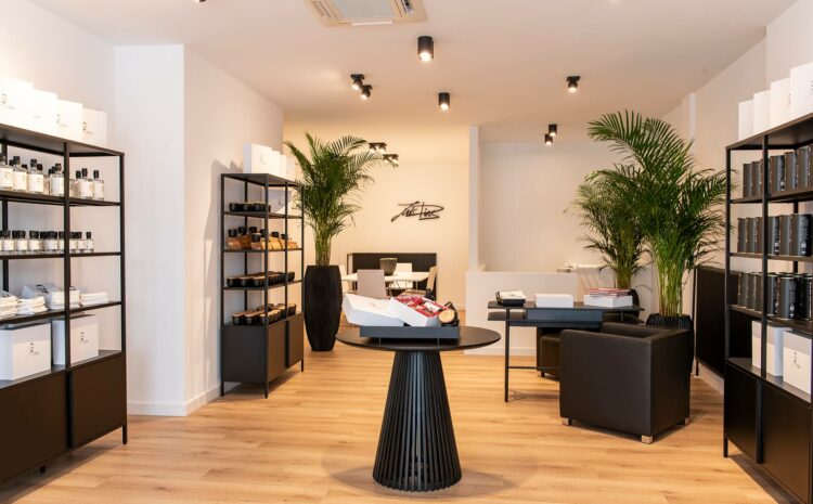 Einladendes Ladenlokal von Casa Luis Dias in Köln, Rodenkirchen, mit prall gefüllten Verkaufsregalen mit hausgemachten Produkten, offenen Geschenkboxen auf einem Tisch in der Mitte, einem gemütlichen Sessel und einem Beratungstisch. Palmen runden das freundliche Ambiente ab.